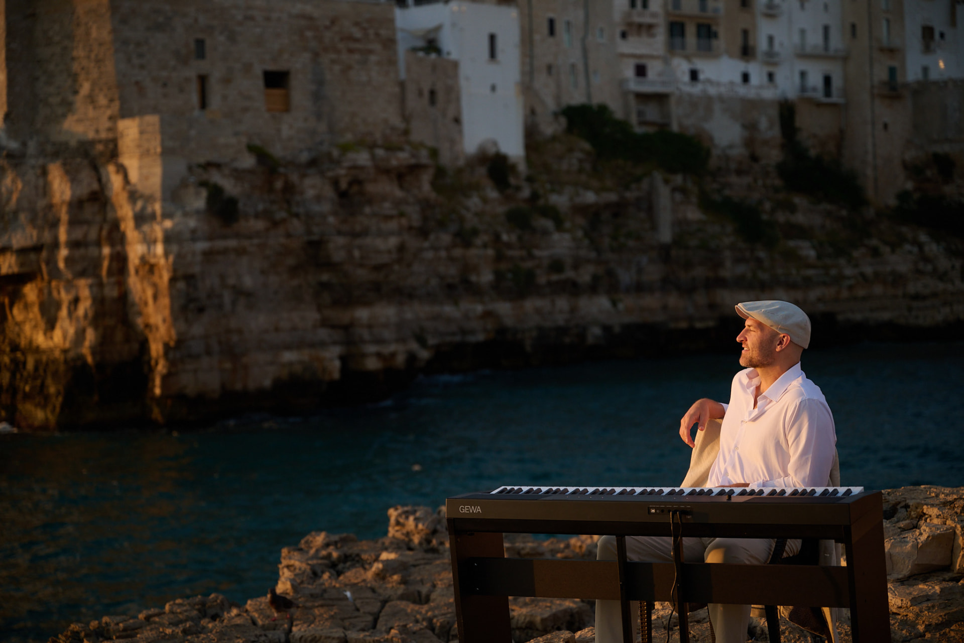 Matthew Mayer playing a portable keyboard on the clifftop rocks at golden hour in Polignano a Mare, Puglia, Italy, with the whitewashed town rising behind him