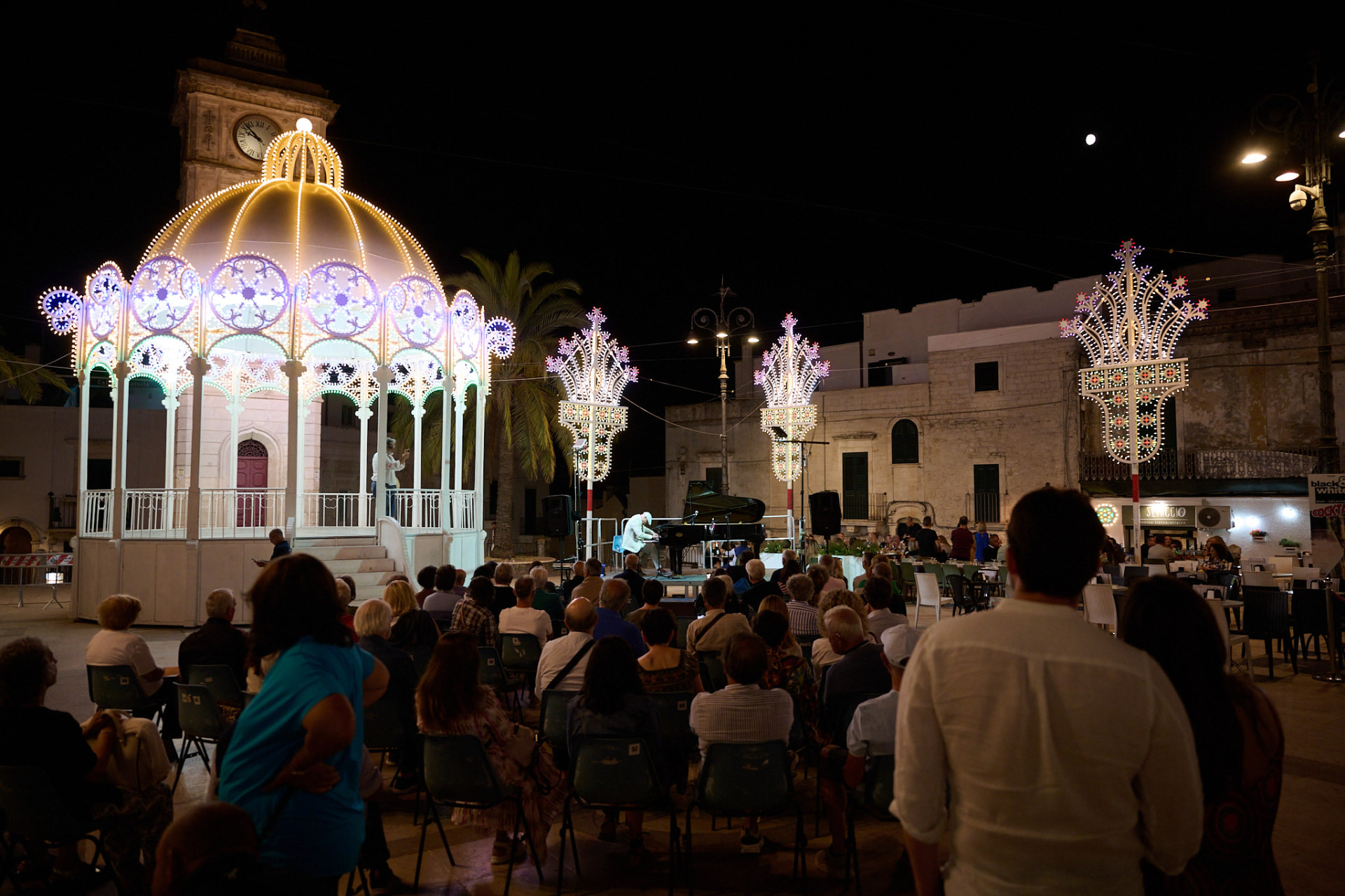 Matthew Mayer performing on a grand piano at night in a festival-lit town square in Ceglie Messapica, Italy, before a seated audience